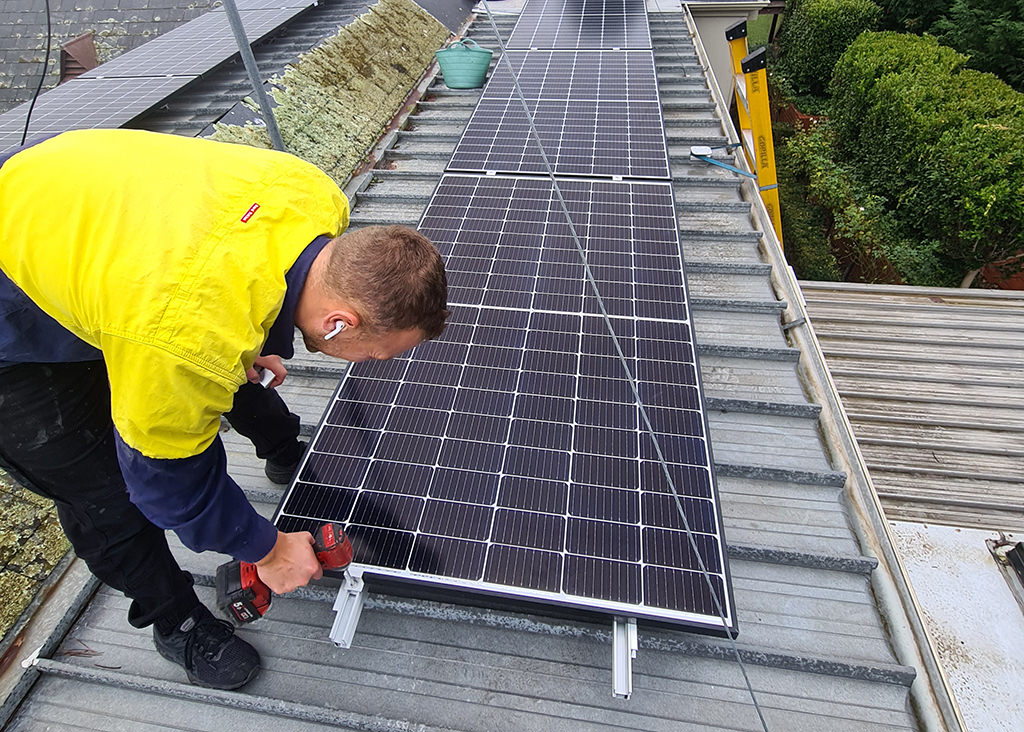 An Australian tradesman in a typical bright top installing a solar panel on a roof.