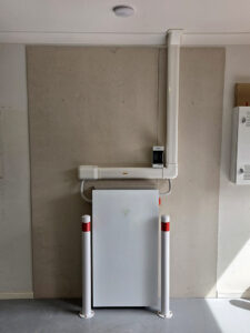Indoor home battery storage unit installed against a wall with protective bollards and surface-mounted conduit above the cabinet
