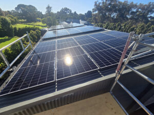 large array of black solar panels newly installed on a grey corrugated metal roof, viewed from scaffolding during a residential installation project.