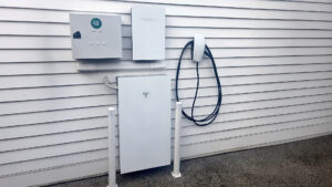 Tesla Powerwall home battery installed outdoors alongside a wall-mounted EV charger, inverter, and protective bollards on a residential building exterior